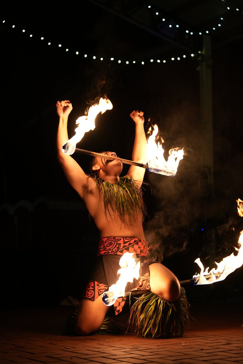 Fire knife dancer kneeling with a flaming staff under string lights